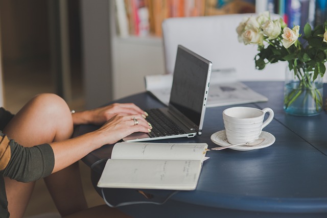 Persoană lucrând la un CV pe laptop, cu o ceașcă de cafea și un jurnal deschis